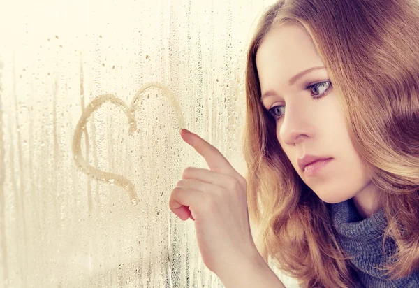 Sad girl draws a heart on the window in the rain — Stock Photo, Image