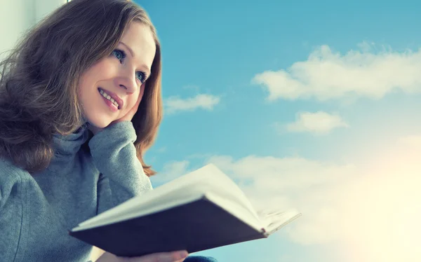 Beautiful young woman reading a book while sitting at a window i — Stock Photo, Image