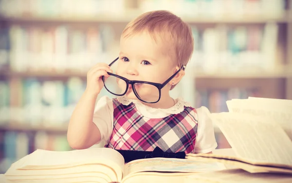 Lustiges kleines Mädchen mit Brille liest ein Buch in einer Bibliothek — Stockfoto