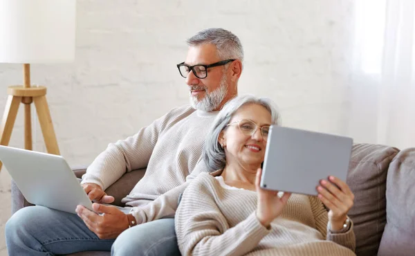 Glad avslappnad gammal pensionerad familj par med hjälp av modern teknik laptop och digital surfplatta hemma — Stockfoto