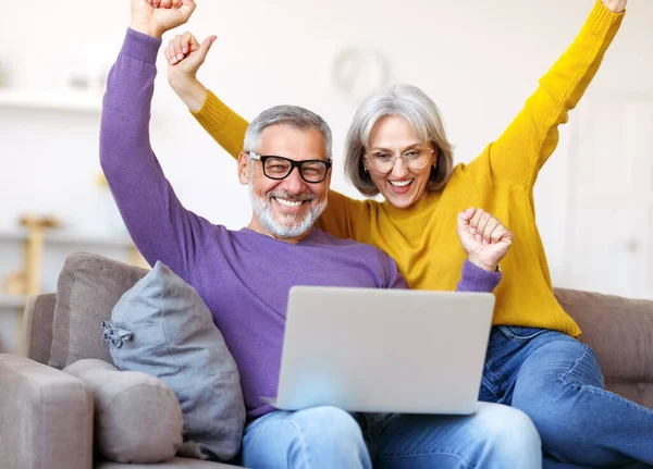 Ενθουσιασμένο ζευγάρι ηλικιωμένων γιορτάζει την επιτυχία, ενώ κάθεται στον καναπέ με φορητό υπολογιστή — Φωτογραφία Αρχείου