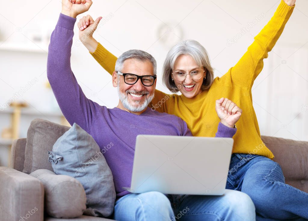 Overjoyed excited senior family couple celebrating success while sitting on sofa with laptop