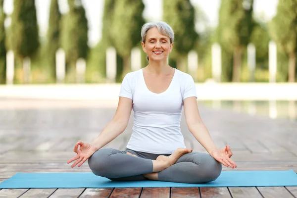 Nyugodt boldog idős nő ül lótuszban pózol matracon reggeli meditáció közben a parkban — Stock Fotó