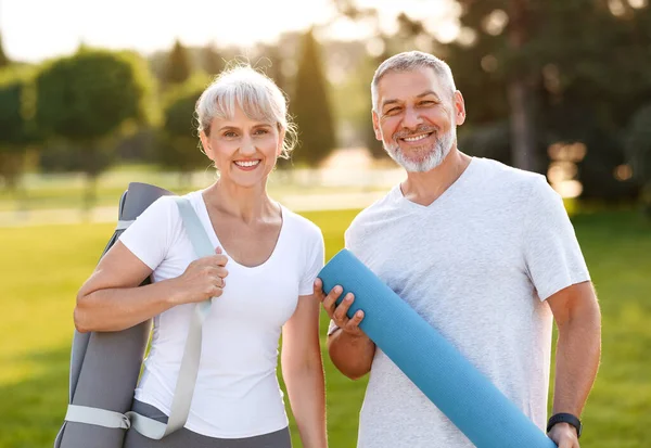 Sonriendo pareja de ancianos marido y mujer abrazando mientras están de pie en el parque con colchonetas de ejercicio — Foto de Stock