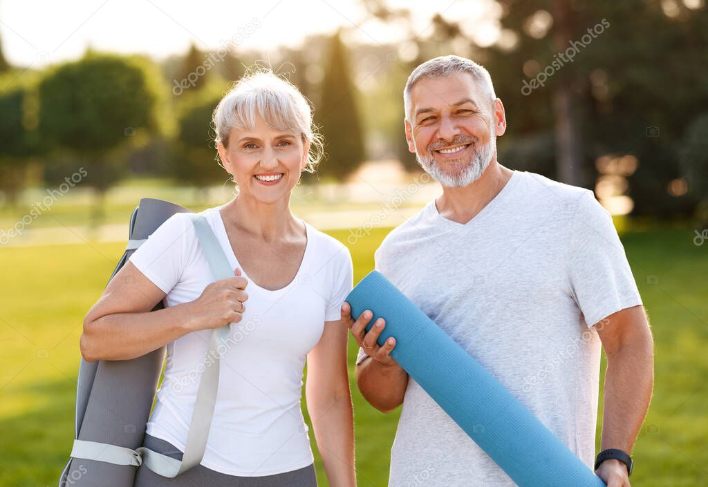 Smiling senior couple husband and wife embracing while standing at park with exercise mats