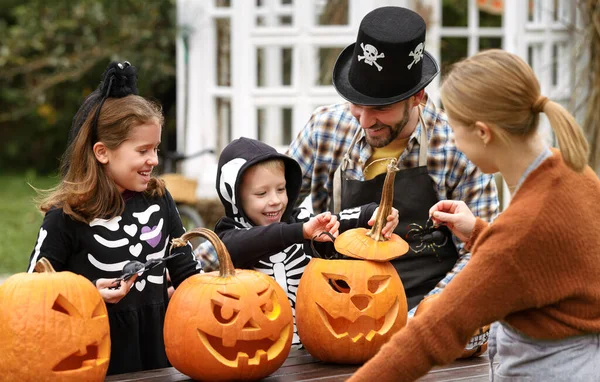 Счастливая молодая семья в костюмах на Хэллоуин вырезает тыквы на заднем дворе — стоковое фото