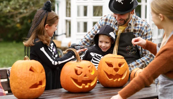 Счастливая молодая семья в костюмах на Хэллоуин вырезает тыквы на заднем дворе — стоковое фото