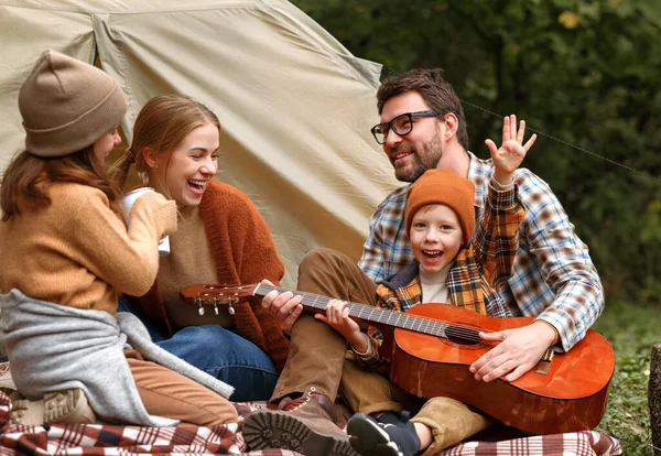 Father, mother and little son sitting near tourist tent and playing guitar during camping