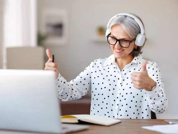 Beautiful smiling mature woman in headphones during online meeting on laptop with collegues at home — Stock Photo, Image