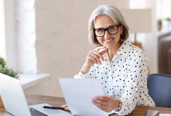 Усміхнена старша жінка тримає паперовий фінансовий документ і дивиться на ноутбук під час роботи з дому — стокове фото