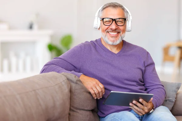 Bonito homem idoso feliz usando tablet digital enquanto relaxa descansando no sofá em casa — Fotografia de Stock