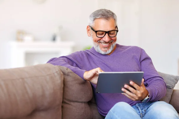 Bonito homem idoso feliz usando tablet digital enquanto relaxa descansando no sofá em casa — Fotografia de Stock