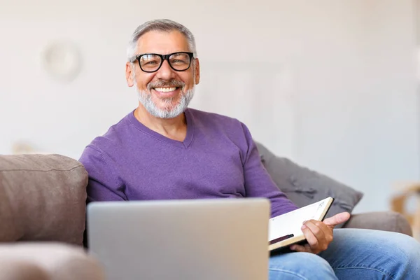 一位英俊快乐的老人，戴着眼镜，坐在沙发上用笔记本电脑远程工作 — 图库照片