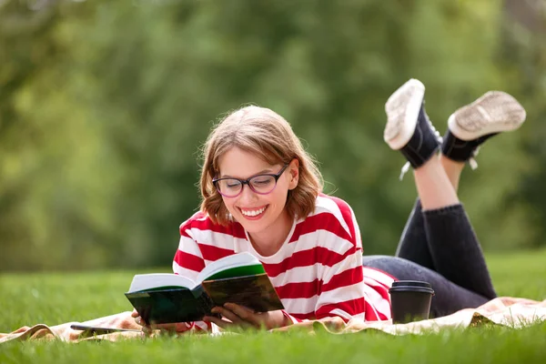 Piękna dziewczyna w okularach z uroczym uśmiechem leżącym na zielonej trawie w parku miejskim z otwartą książką — Zdjęcie stockowe