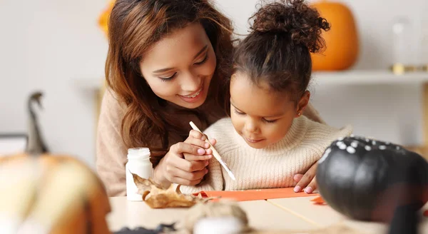 Cute little african american girl painting Halloween pumpkin with mom together at home — Stock Photo, Image