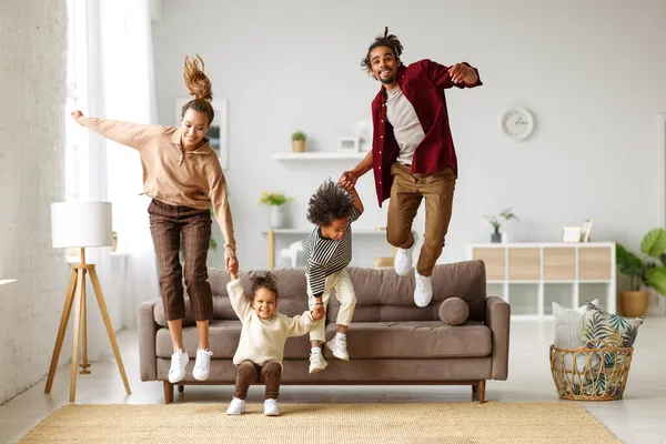 Aktiva unga afrikanska amerikanska föräldrar hoppar från soffa till golv med två små barn hemma — Stockfoto