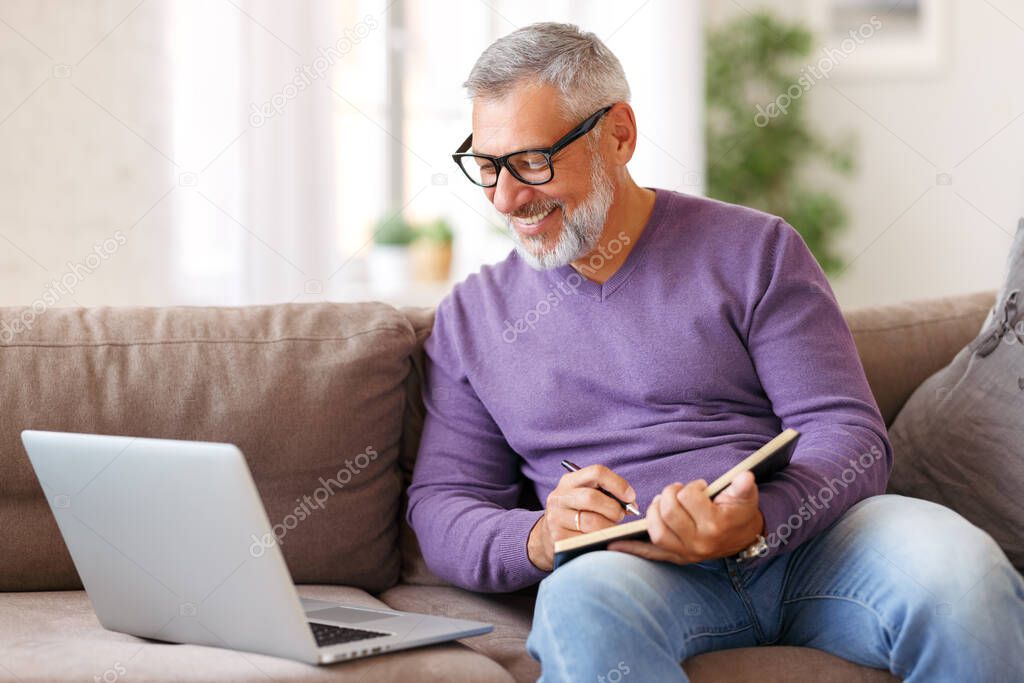 Handsome happy senior man in glasses working remotely while sitting on sofa with laptop computer