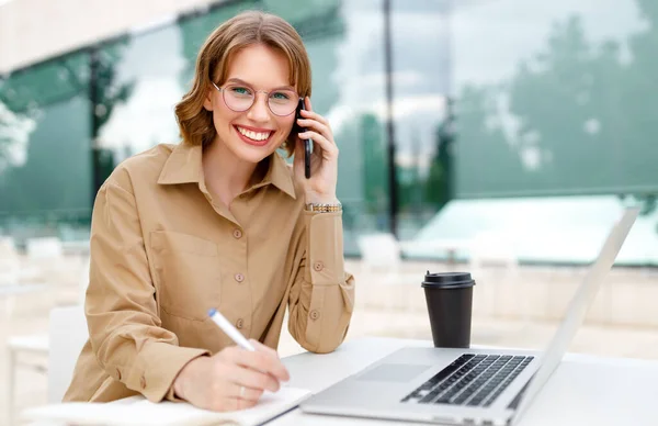 Feliz joven asistente de oficina trabajando remotamente en línea sentado con el ordenador portátil y café fuera —  Fotos de Stock