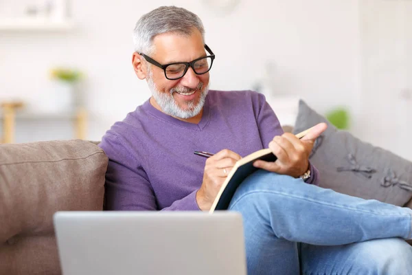 Guapo feliz hombre mayor en gafas de trabajo a distancia mientras está sentado en el sofá con el ordenador portátil —  Fotos de Stock