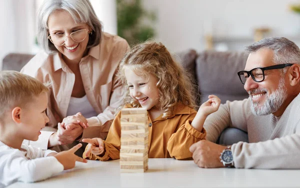 Возбужденные дети играют в игру Дженга дома с позитивными старшими бабушкой и дедушкой, сидя на диване — стоковое фото