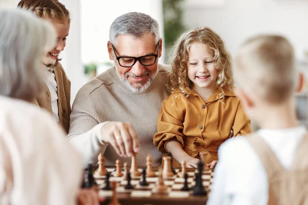 Çocuklar, abiler ve ablalar büyük anne ve babalarıyla oturma odasında otururken satranç oynuyorlar.