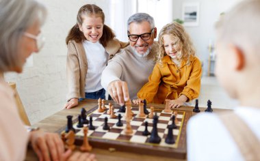 Çocuklar, abiler ve ablalar büyük anne ve babalarıyla oturma odasında otururken satranç oynuyorlar.