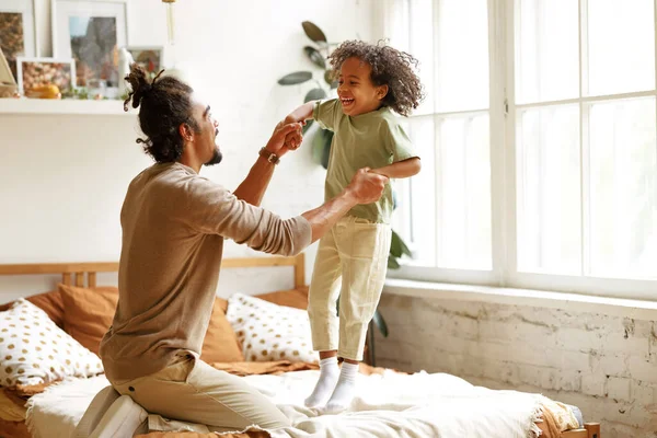 Mutlu Afro-Amerikan baba ve oğlu evde oynarken yatakta zıplayıp gülüyorlar. — Stok fotoğraf