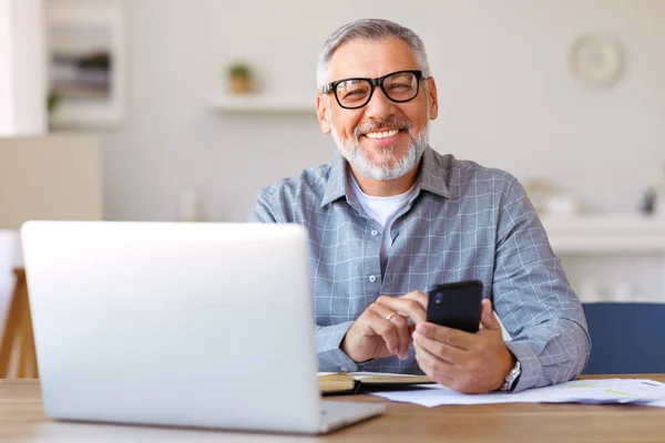 Hombre mayor sonriente usando el teléfono móvil mientras está sentado en su acogedor lugar de trabajo con el ordenador portátil en casa — Foto de Stock
