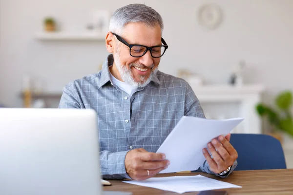 Guapo sonriente hombre mayor leyendo documentos financieros mientras trabaja remotamente en el ordenador portátil en casa —  Fotos de Stock