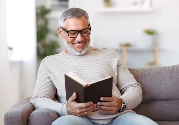 Šťastný starší muž s šedivými vlasy v brýlích čtení knihy doma, odpočívá v obývacím pokoji — Stock fotografie