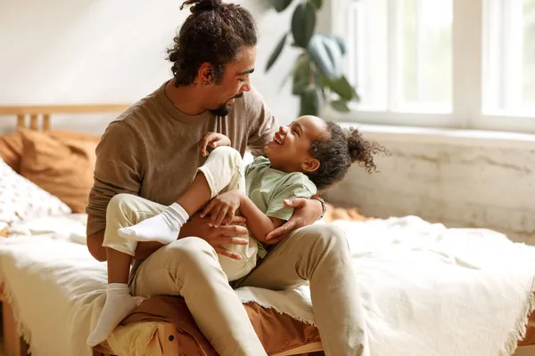 Happy afro american family father and son laughing and playing while sitting on bed at home