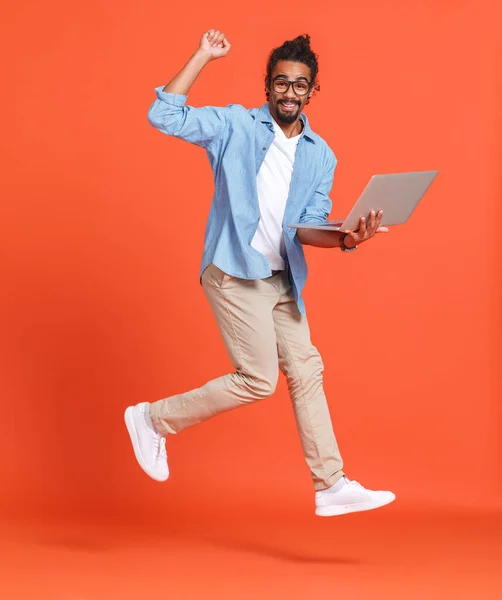 Joven feliz hombre afroamericano alegre con computadora portátil saltando sobre fondo naranja —  Fotos de Stock