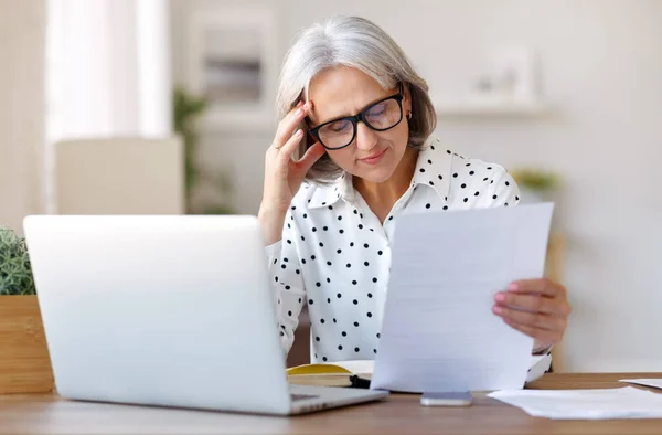 Hermosa mujer mayor sonriente leyendo documentos financieros mientras trabaja remotamente en el ordenador portátil en casa —  Fotos de Stock