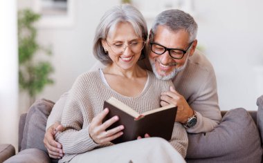 Mutlu gülümseyen son sınıf aile çifti evde birlikte kitap okuyor.