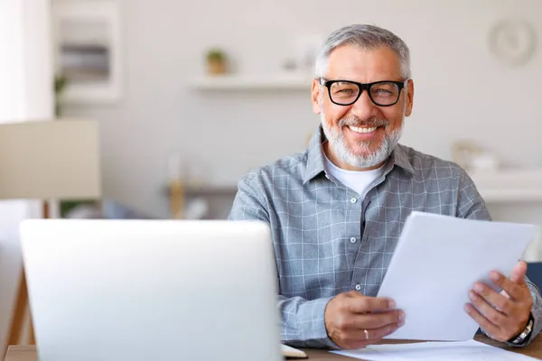 . Knappe glimlachende oudere man die financiële documenten leest terwijl hij thuis op afstand op een laptop werkt — Stockfoto
