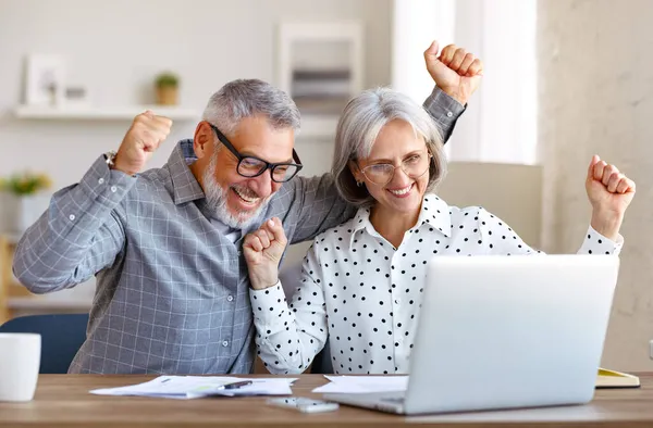 Happy senior couple celebrating success while sitting at table with open laptop at home — Stock Photo, Image