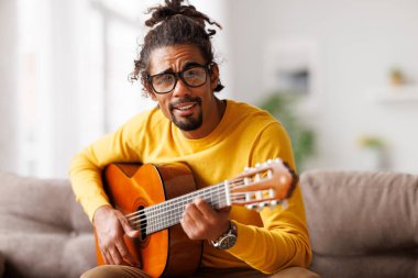 Genç, neşeli Afrikalı Amerikalı adam evde akustik gitar çalıyor, oturma odasında kanepede oturuyor.
