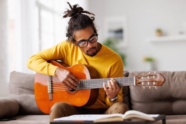 Genç, neşeli Afrikalı Amerikalı adam evde akustik gitar çalıyor, oturma odasında kanepede oturuyor.