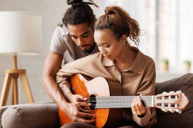 Hafta sonları evde dinlenirken kanepede oturup erkek arkadaşına gitar çalan melez bir kadın.