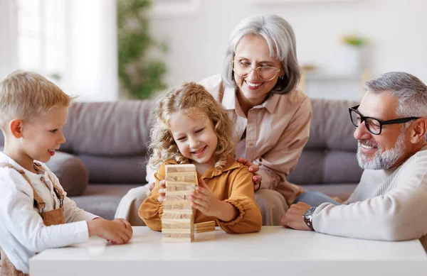 Ενθουσιασμένα παιδιά παίζουν το παιχνίδι Jenga στο σπίτι με θετικά ηλικιωμένους παππούδες, ενώ κάθεται στον καναπέ — Φωτογραφία Αρχείου