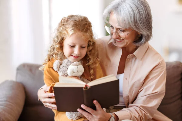 Cute little girl granddaughter reading book with positive senior grandmother — Stock Photo, Image