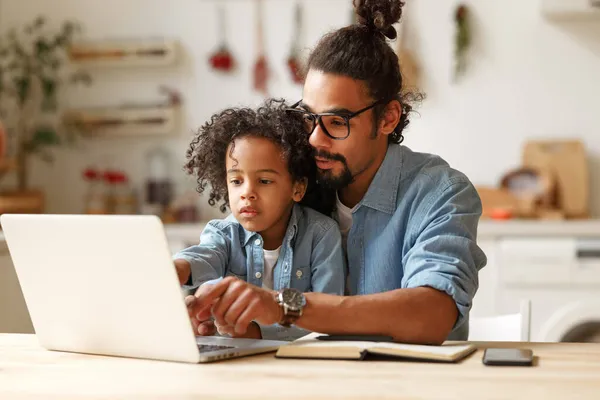 En ung afroamerikansk far som hjelper skolegutt med lekser på laptop under fjernundervisning – stockfoto