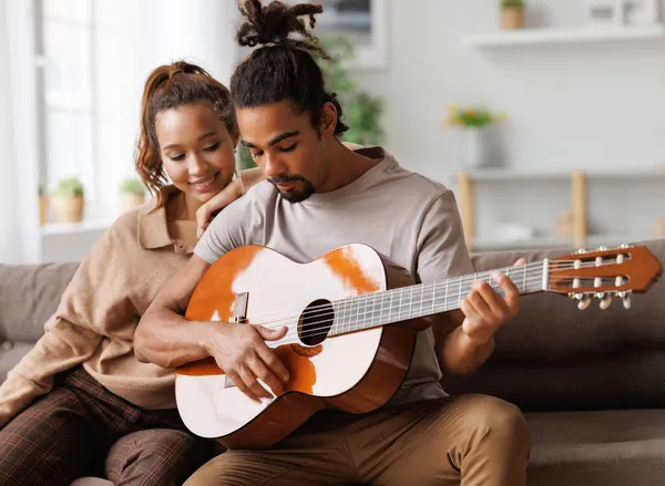 Молодий усміхнений афроамериканський музикант грає інструментальну музику на гітарі для щасливої дівчини — стокове фото