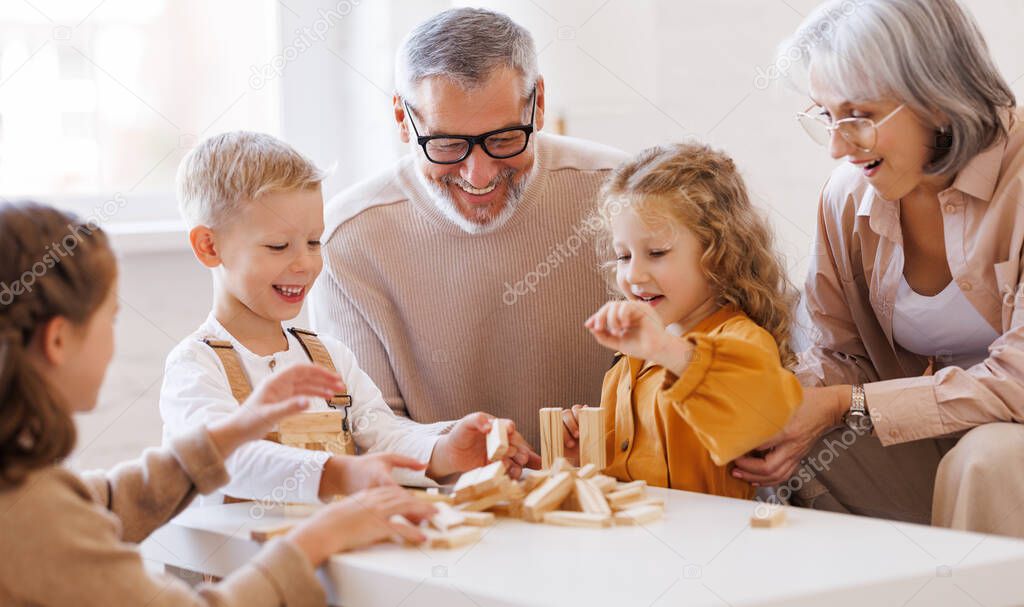 Excited children playing game Jenga at home with positive senior grandparents while sitting on sofa
