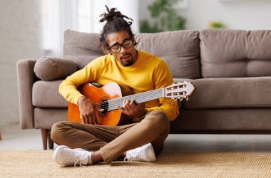 Genç Afro-Amerikalı adam evde akustik gitar çalıyor, oturma odasında yerde oturuyor.