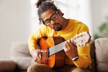 Genç, neşeli Afrikalı Amerikalı adam evde akustik gitar çalıyor, oturma odasında kanepede oturuyor.