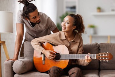 Hafta sonları evde dinlenirken kanepede oturup erkek arkadaşına gitar çalan melez bir kadın.