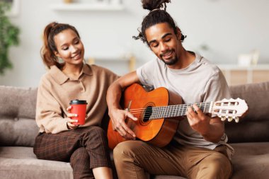 Genç gülümseyen Afrikalı Amerikalı müzisyen mutlu kız arkadaş için gitarda enstrümantal müzik çalıyor.