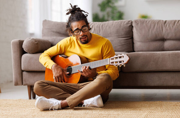 Young joyful african american man playing acoustic guitar at home, sitting on floor in living room