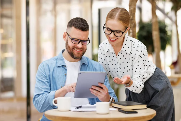 Two positive coworkers man and woman chatting during coffee break in cafe, looking at digital tablet — Stock Photo, Image